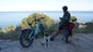 Zwei Himiway D5 Fatbikes in Blau und Schwarz auf einem Berg mit Meerblick, ein Mann lehnt am schwarzen Fatbike, neben ihm sitzt ein Hund.